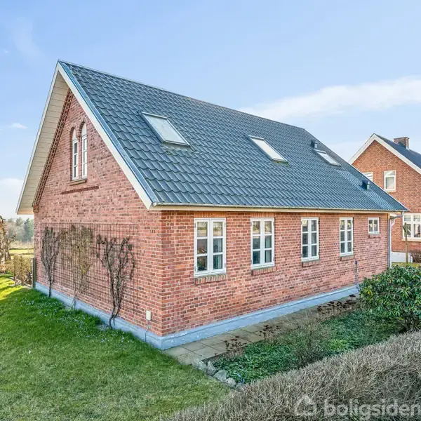 Et murstenshus med sort tag har flere vinduer og kviste. Det står i en velplejet have med tilstødende græsplæne og buske. Gårdhave i baggrunden, klart himmel.