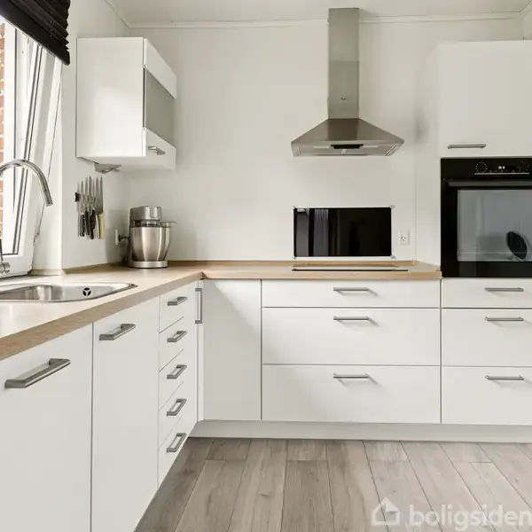 Et moderne køkken har hvide skabe og stålgreb. Der står en ovn, mikroovn og blender på bordet. Rummet er lyst med store vinduer og en udsigt til grønt udenfor.