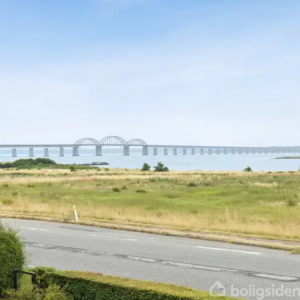 En bro strækker sig over vandet i det fjerne, mens en vej og frodige grønne områder omgiver landskabet i forgrunden under en klar, blå himmel.