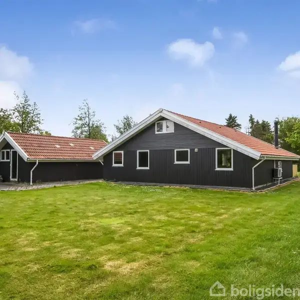 Sort hus med rødt tag står stille i en grøn have omkranset af træer og buske under en blå himmel med spredte skyer.