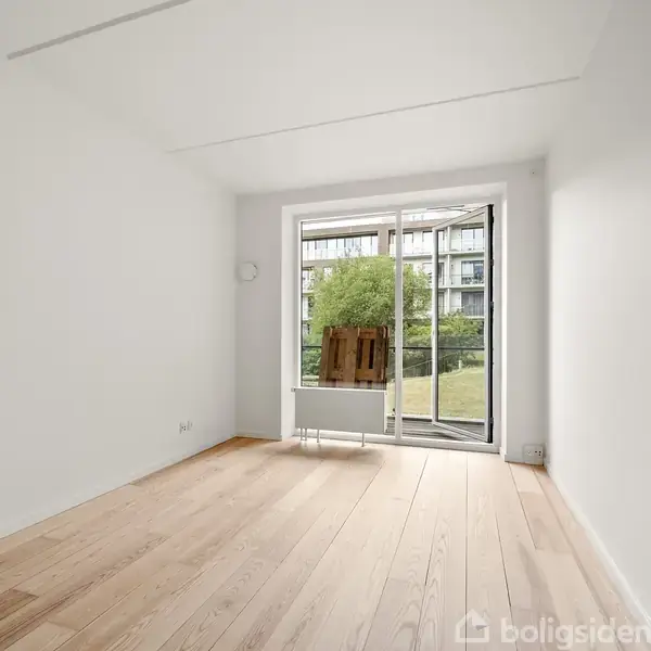 Et tomt rum med trægulv og hvide vægge har en åben dør til en balkon. Udenfor ses græs og en bygning i baggrunden.
