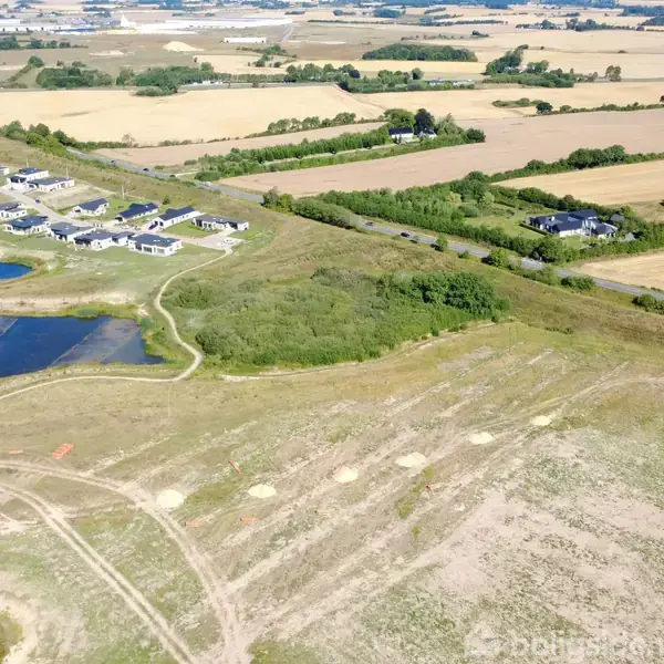 Boligområde med rækkehuse ligger ved siden af små søer og marker. Der er åbne marker og skove i baggrunden, som skaber et fredfyldt landligt miljø.
