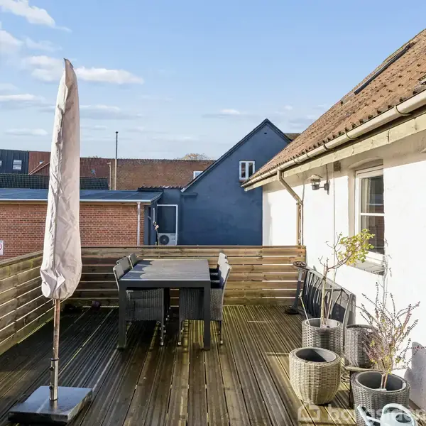 En træterrasse har et bord med fire sorte stole og en sammenfoldet parasol. Terrassen er omgivet af et træværk og tilstøder et hvidt hus med skråtag.