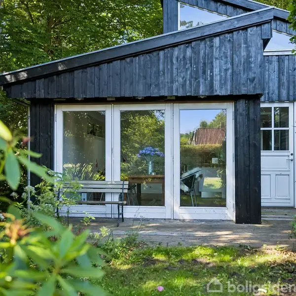 Et sort træhus med store vinduespartier står i en grøn have med frodige buske. En hvid dør er synlig, og der er et bord og en bænk foran huset.
