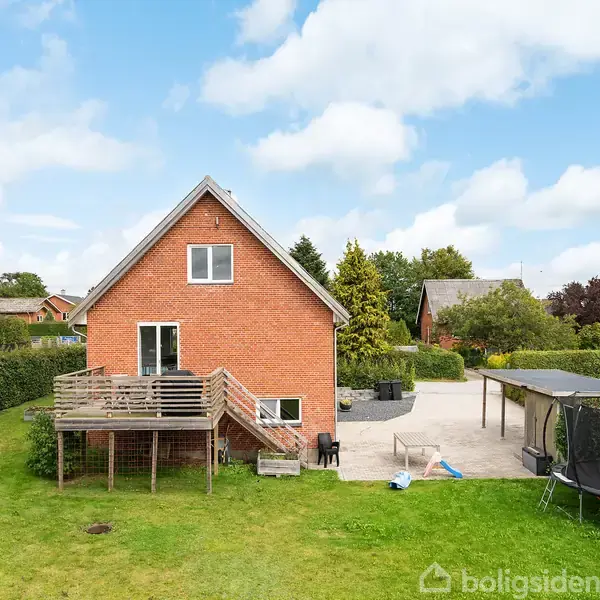 Rødt murstenshus med træterrasse, placeret i en have med græsplæne. Til højre ses en trampolin og flisebelagt terrasseområde, omgivet af hække og nærliggende huse.