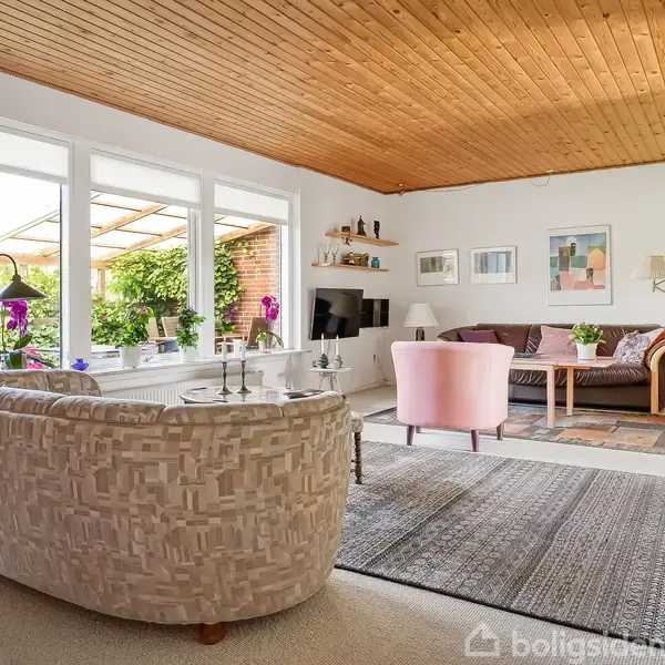 Stue med møbler som sofa, lænestole og sofabord, der står i en lys indretning med trægulv og loft. Store vinduer viser udsigt til en grøn have. Ingen tekst fundet.