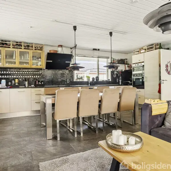 Køkken med træspisebord og seks stole i en moderne, lys indretning. Indbyggede skabe og ovne er i baggrunden, mens stuen vises med sofa og sofabord i forgrunden.