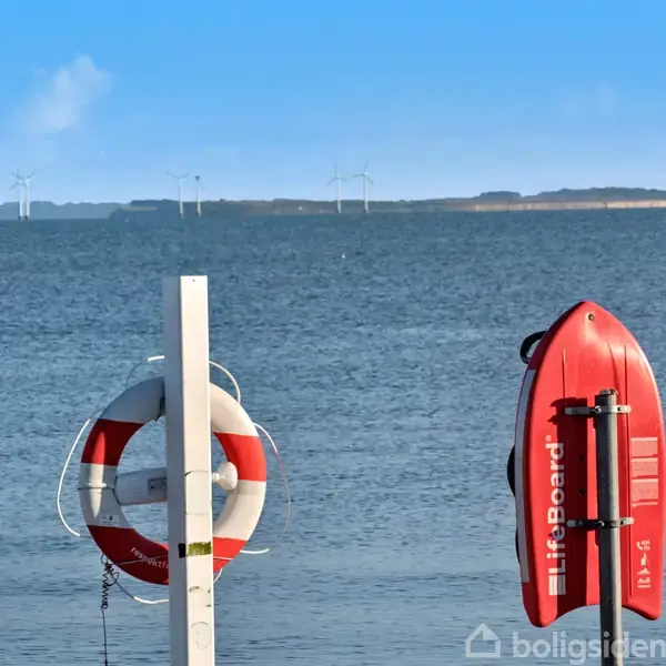 Redningskrans og rescue board hænger på stativer ved kysten. I baggrunden ses havmøller og hav. Tekst på rescue board: "Lifeboard".