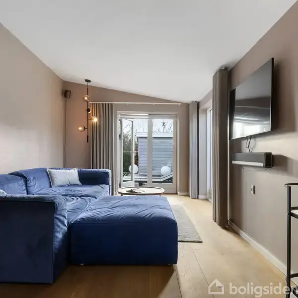 Blå sofa står i stuen, vendt mod en vægmonteret fladskærm. Bag sofaen fører glasdøre ud til en terrasse. Rummet er lyst og moderne indrettet med trægulv.