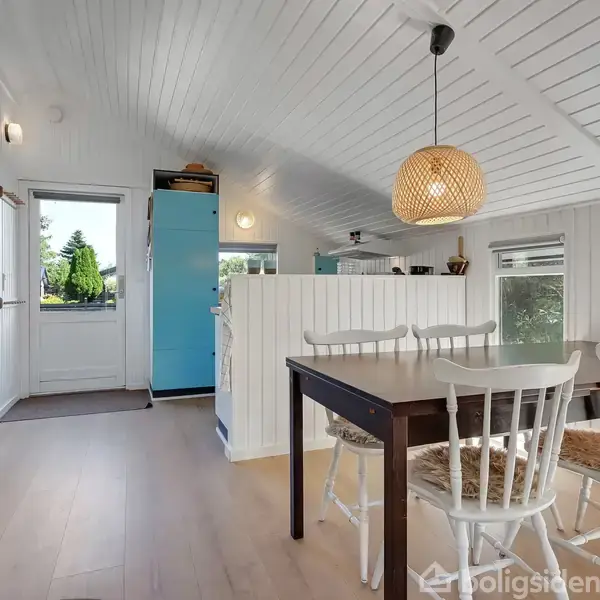Spisebord med fire stole står i et lyst rum, oplyst af en hængende lampe. I baggrunden ses køkkenet med et blåt skab. En brændeovn opvarmer rummet.