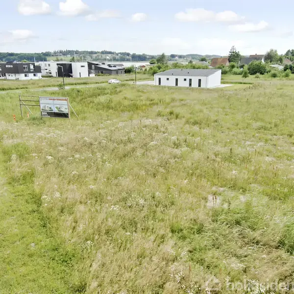 En åben græsmark med en skilt ved siden af, omgivet af moderne bygninger i baggrunden på en solskinsdag. Skiltet har teksten: "Attraktive byggegrunde i Nyrup Bakker. 30% solgt. Kontakt os på 70108099".