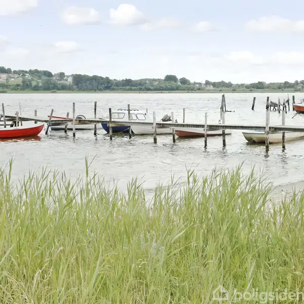 Både ligger fortøjet ved en træbro på en sø. De er omgivet af vand og græs i forgrunden med en grøn kystlinje og bebyggelse i baggrunden.