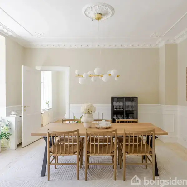 En træspisebord omgivet af seks stole står i et lyst, elegant spisestue med hvid ornamenteret loft, lyse vægge og en moderne lysekrone. Et skab ses i baggrunden, og rummet har planter ved døren.