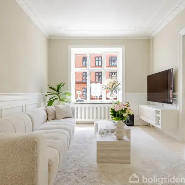 En lys stue har en hvid sofa, et sofabord med planter, en vægmonteret fladskærm og et stort vindue, der giver udsigt til en rødstensbygning udenfor.