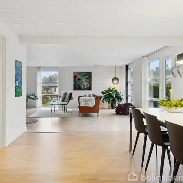 Et stilfuldt, lyst rum med trægulv har en spisestue med sorte stole og et bord. En stue med stole og planter ses i baggrunden. Lyse vægge og store vinduer skaber luftighed.