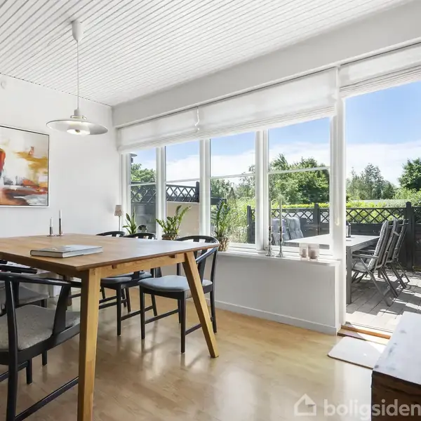 Spisebord med sorte stole står i et lyst rum med trægulv. Store vinduer og en åben dør leder ud til en terrasse. Kunstmaleri hænger på væggen.