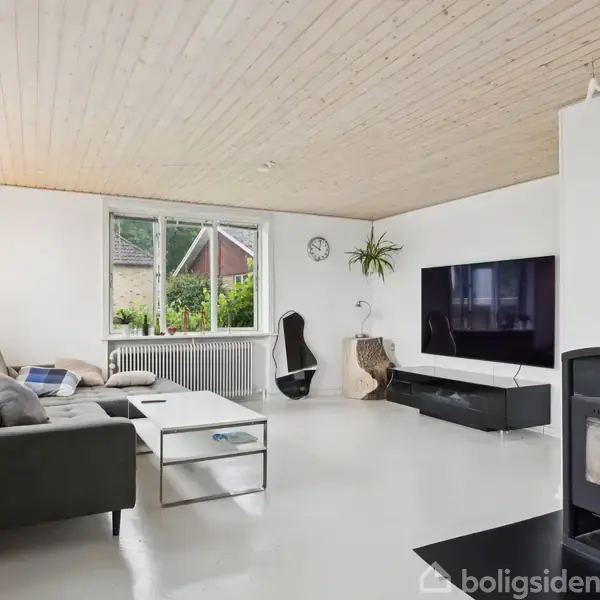 En stue med en grå sofa står ved et stort vindue. En pejs brænder til højre. En fladskærms-TV hænger på væggen. Indretningen er lys og moderne.