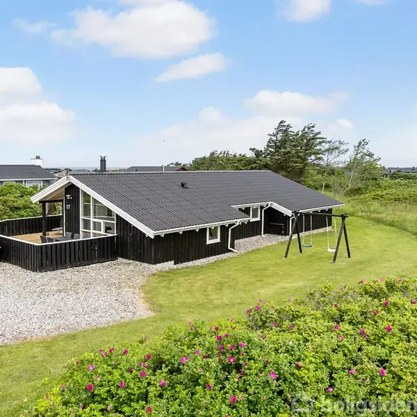 Et sort træhus står på en grusvej med en terrasse, omkranset af en velholdt græsplæne og buske. En enkelt gynge er placeret ved siden af huset under en blå himmel.