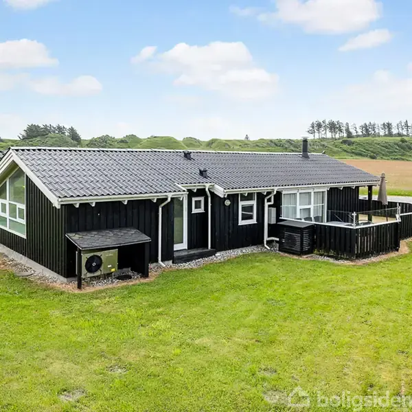 Sort træhus står stille på en grøn græsplæne omgivet af åbne marker og lave bakker under en blå skyet himmel. Der er en mindre terrasse foran huset.