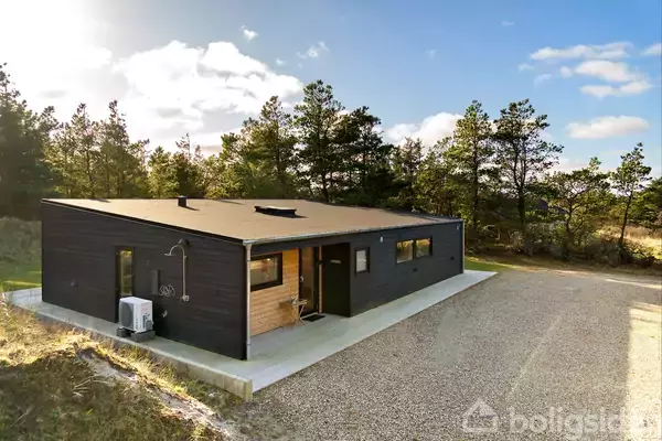 Et sort, rektangulært hus står stille på en grusvej omringet af grønne træer og græsarealer. Huset har store vinduer og en træterrasse.