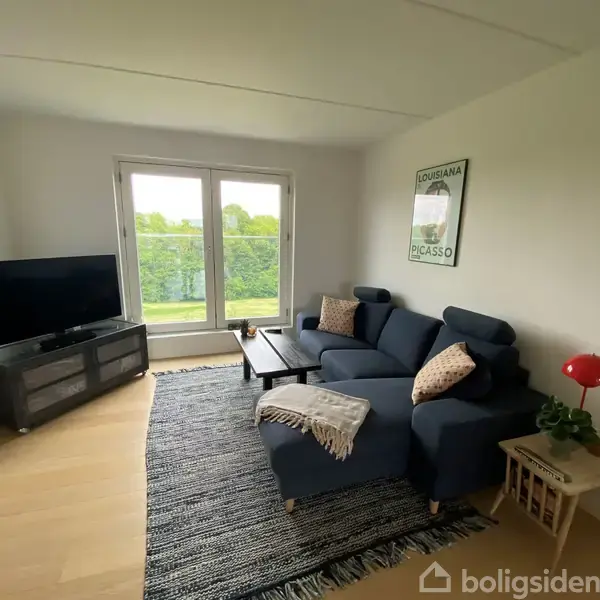 En stue indeholder en blå sofa med puder, en træbord med en lampe, og et fjernsyn på et skab. Store vinduer viser grønne træer udenfor. Vægkunst siger "LOUISIANA PICASSO".