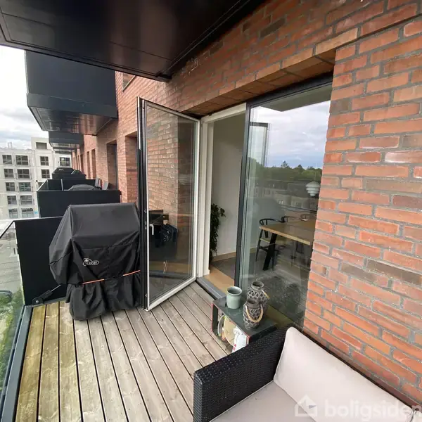 Altan med træterrasse og glasrækværk, hvor en gasgrill står dækket til. En dør åbner ind til en stue med planter. Høje bygninger ses i baggrunden.
