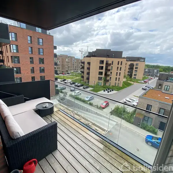 Balkon møbleret med sofa og bord; set fra en højt beliggende lejlighed. Omgivet af moderne bygninger, parkeringsplads og grønne områder. Skyerne dækker delvis himlen.