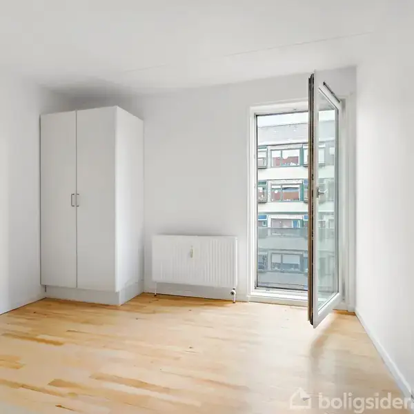 Et tomt værelse med hvidt skab står på trægulvet, en radiator under et åbent vindue, med udsigt til en bymæssig baggrund.