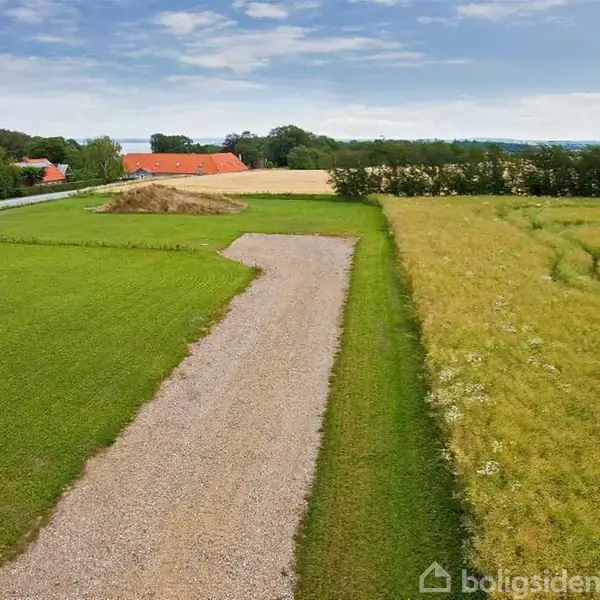 Grusvej strækker sig gennem landområde, omgivet af grønne marker og skov i baggrunden. Til venstre ses røde tage fra enkelte huse langs vejen.