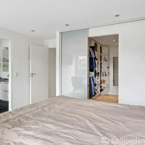 Soveværelse med stor seng i forgrunden. Skydedør fører til en walk-in garderobe. Til venstre er der en åbnet dør til et køkken med sorte fliser.
