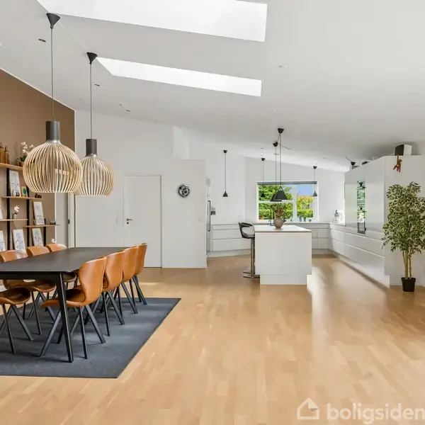 Spisebord med træstole står på et tæppe, omgivet af moderne opholdsrum med trælaminatgulv. Over bordet hænger store lamper. Planter og dekorative elementer skaber en hyggelig atmosfære.