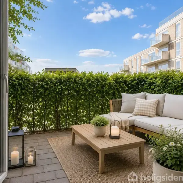 Sofa med puder står på altan. Et lavt bord bærer lys og planter. Omkranset af grøn hæk. Moderne lejlighedsbygninger kan ses i baggrunden under blå himmel.