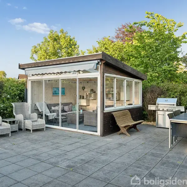 Et moderne udestue med glasvægge står på en flisebelagt terrasse. Der er havemøbler og en grill, omgivet af frodig vegetation og blå himmel. Markisen er rullet ud.
