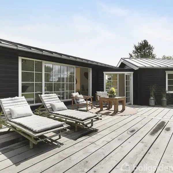 Terrasse med to liggestole står i solen foran et sort træhus med store vinduespartier. Et havebord med stol findes ved siden af. Grøn græsplæne omgiver terrassen.
