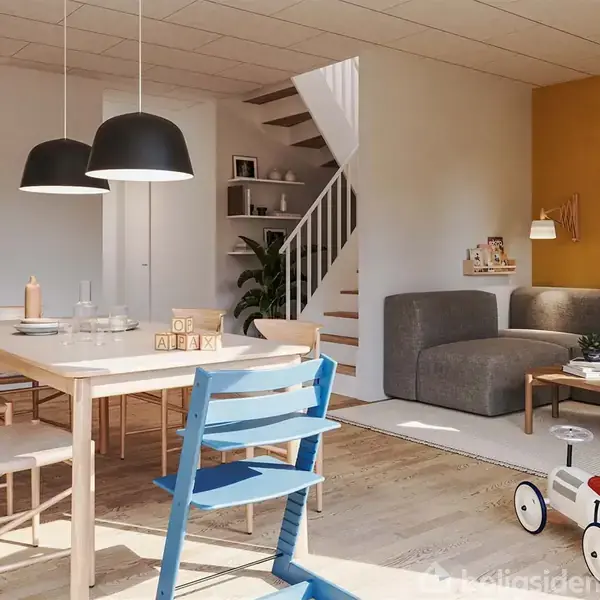 En moderne spisestue med træbord, stole og en blå barnestol. Stuen har en grå sofa, tæppe og legetøjsbil. På væggen hænger kunst, trappen fører ovenpå. Hyggelig atmosfære.