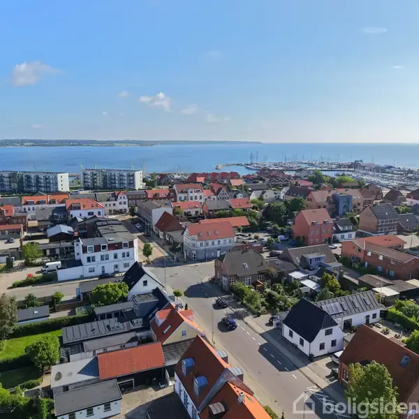 Byområde med røde tage og bygninger, der spreder sig ud mod en kystlinje. I baggrunden ses havet med en marina til højre og skovklædte områder i det fjerne.