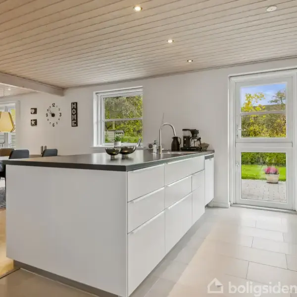 Et moderne køkken med hvid køkkenø er centralt placeret. Døre og vinduer åbner mod en grøn have. Spisebord med stole står i baggrunden.