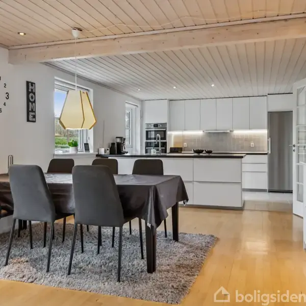 Spisestue med seks stole står omkring et bord dækket med en dug; nær et åbent, moderne køkken med hvide skabe og grå bordplader. Væggen har tekst: "HOME". Korkgulv og lyse vægge.