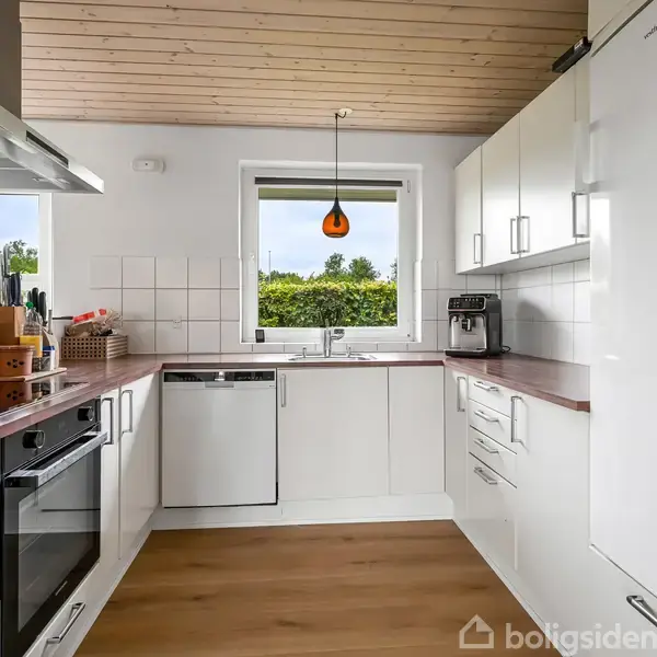 Et moderne, hvidt køkken med indbyggede skabe og apparater, herunder ovn og opvaskemaskine, mens en kaffemaskine står på bordet. En orange lampe hænger over vasken ved vinduet.