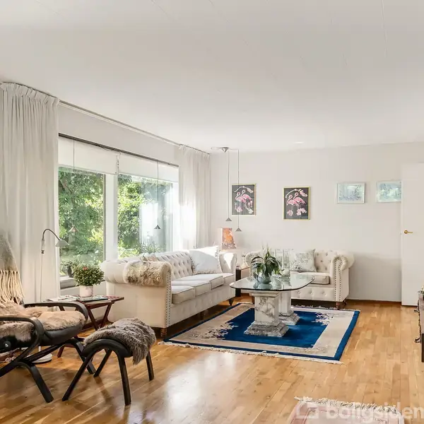 Stue med sofa og stol betrukket med lyse mønstrede tæpper, placeret ved vinduer. Træbord med dekorative planter på tæppe, omgivet af kunstværker på væggene. Planter og møbler skaber en hyggelig atmosfære.