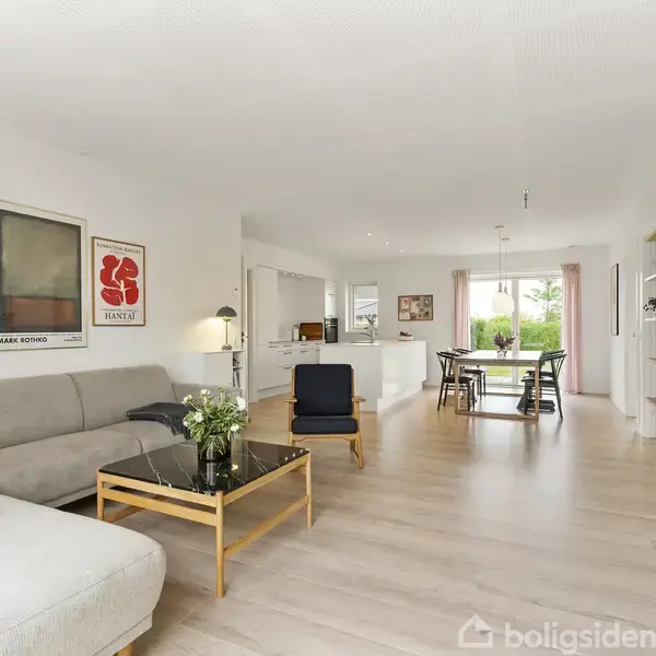Grå sofa med træbord og grøn plante i stuen. Plakater hænger på væggen. Stue-alrum rummer spiseområde og lysindstrømning gennem vinduer. Trægulv og åbent layout skaber en rummelig atmosfære.