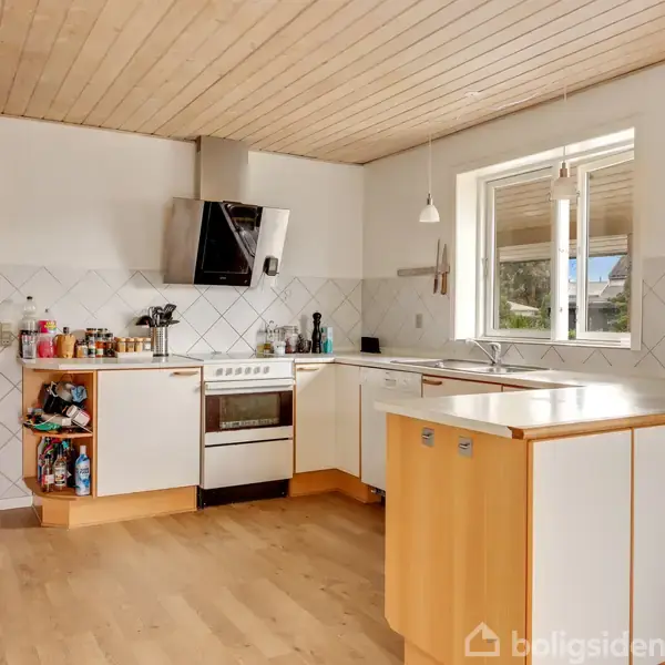 Køkken med hvide skabe står tomt, mens dagslys strømmer ind gennem vinduet, afslørende en komfur med emhætte og redskaber på bordpladen i en skandinavisk indstilling.