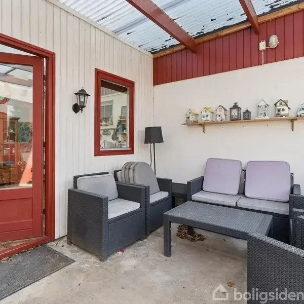 En terrasse med sorte kurvemøbler, herunder sofa og stole, omkring et bord. På væggen er der små dekorative fuglehuse. Terrassen har en glasdør med udsigt til et indendørs rum.