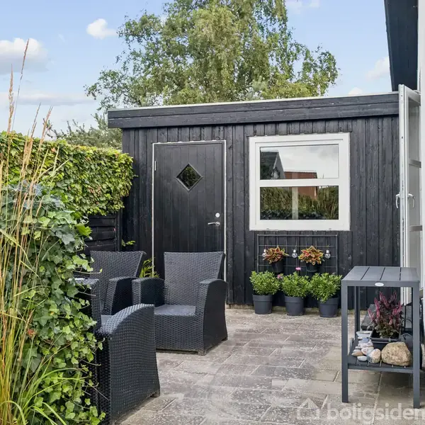 Sort skur med en dør og et vindue står ved en flisebelagt terrasse, omkranset af grønne planter. Lænestole er placeret til afslapning, og en siddeplads med dekorative genstande ses.