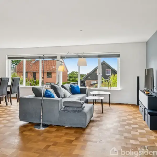 Grå sofa med blå puder står foran vinduer, ved siden af tv og brændeovn. Spisebord med stole er i baggrunden. På væggen står: "WALK ON WALK ON WITH HOPE IN YOUR HEART AND YOU'LL NEVER WALK ALONE."