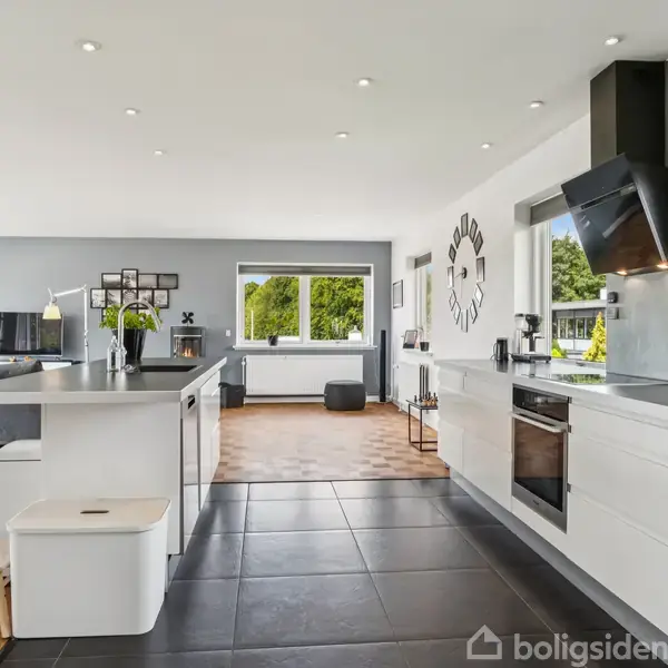 Køkken med moderne indretning har hvidt skab, sort flisegulv, og en stor køkkenø med taburet. I baggrunden er en åben stue med spiseplads, sofa og store vinduer mod grøn udsigt.