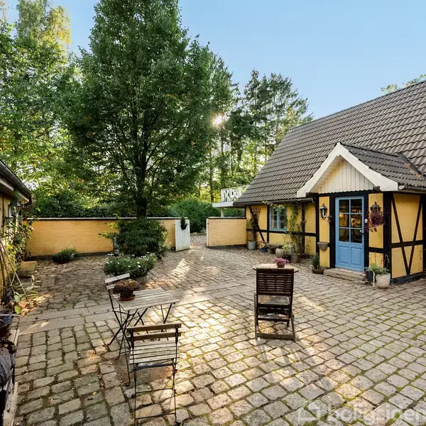 Et bindingsværkshus med blå døre og vinduer står i en gårdsplads med brosten. To træstole og et bord står foran huset. Omgivet af grønne træer.