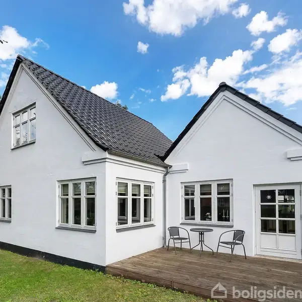 Et hvidt hus med mørkt tag står stille. Foran huset er en terrasse med bord og stole. Græsplæne og buske omgiver huset under en klar blå himmel.