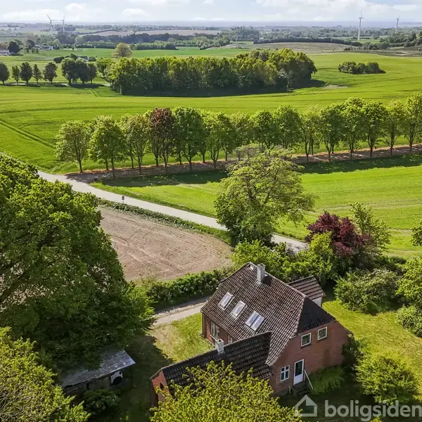 Et rødt murstenshus står omgivet af frodige træer og grønne marker. Et snoet landvej skærer gennem det åbne landskab, der strækker sig under en delvist skyet blå himmel.