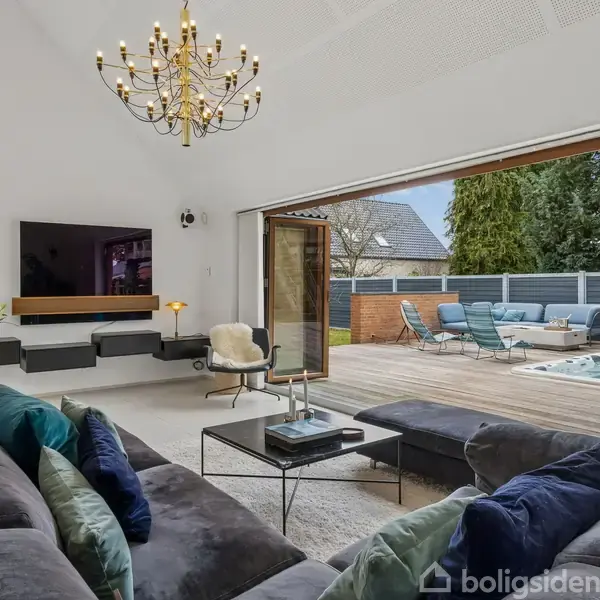 En moderne stue med en stor, mørk sofa og farverige puder vender mod et tv på væggen. Rummet åbner ud til en terrasse med lænestole og et spabad i en have.